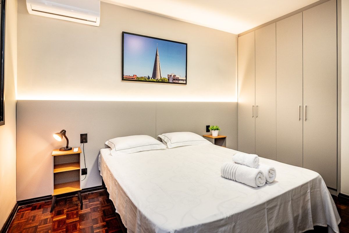 Hotellihuone, jossa on sänky, yöpöytä, seinälle asennettu ilmastointilaite, kehystetty taulu ja vaatekaappi.