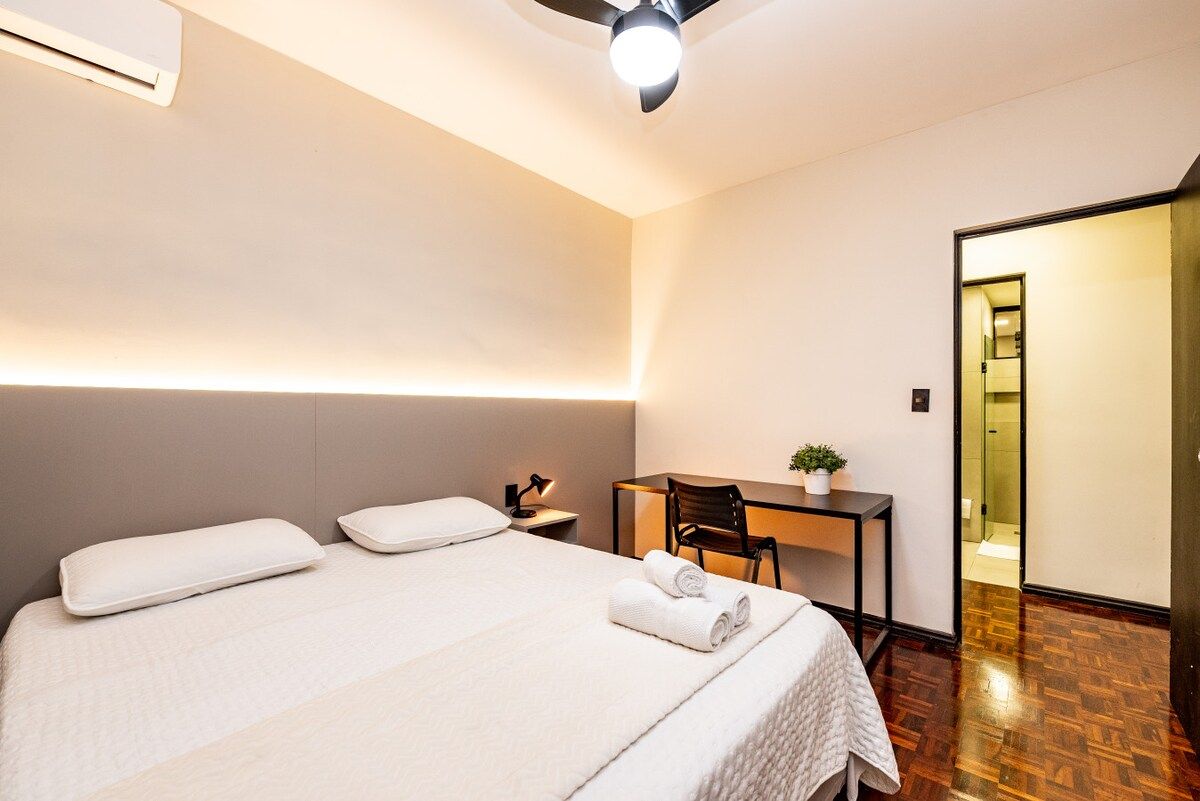Quarto de hotel com cama, escrivaninha, cadeira e uma pequena planta.