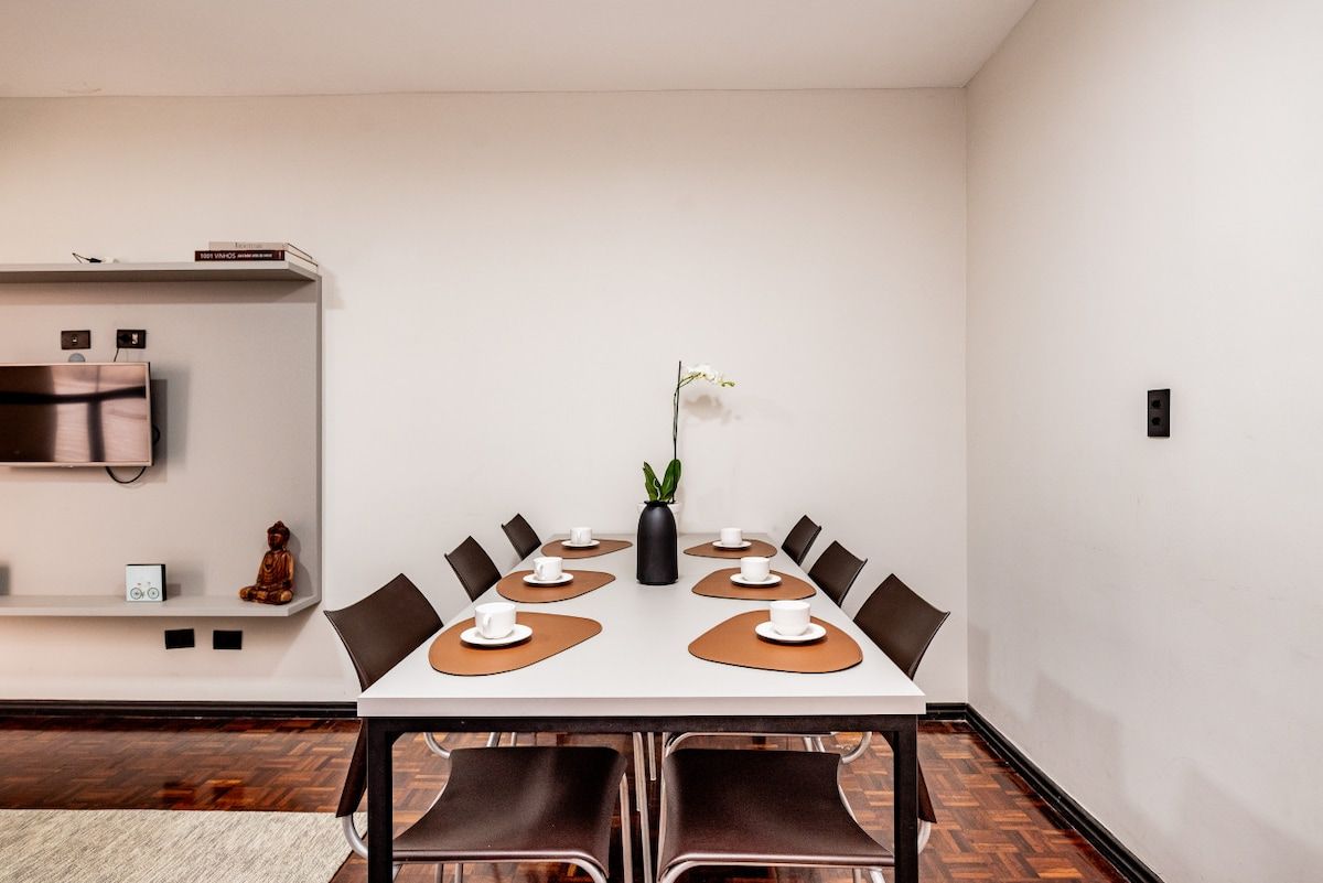 Sala de conferências com uma mesa branca, jogos americanos marrons e cadeiras pretas.