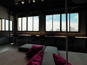 A modern hotel room with a large window, a desk, and a sofa with cushions.