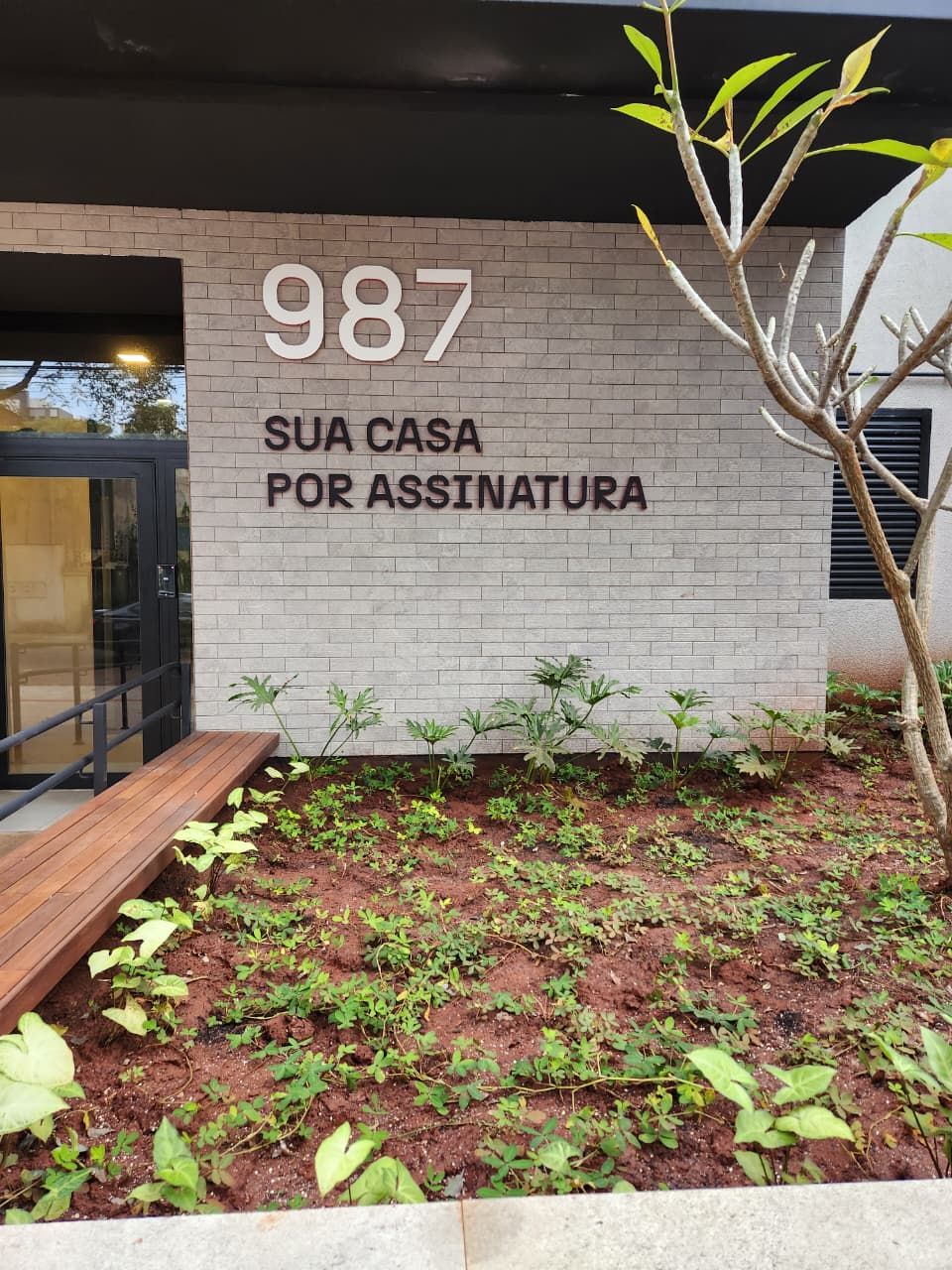 Un edificio con un letrero que dice '987 SUA CASA POR ASSINATURA' y una pequeña área de jardín con plantas.
