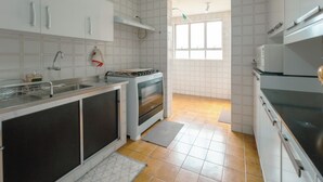 Cozinha com armários brancos, fogão, micro-ondas e janela que traz luz natural.