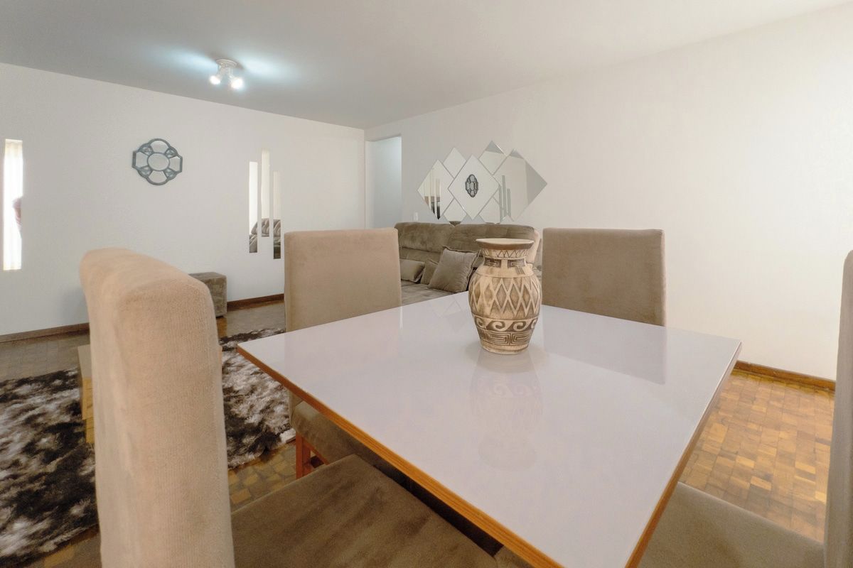 Un comedor con una mesa blanca, cuatro sillas, un jarrón decorativo y una alfombra estampada.