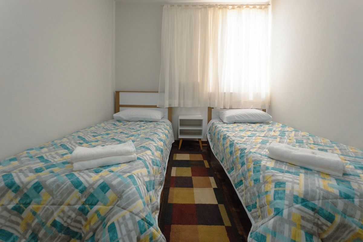 Ein Zimmer mit zwei Einzelbetten, einem kleinen Nachttisch, einem Beistelltisch und einem Fenster mit durchsichtigen Vorhängen.