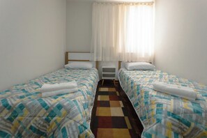 A room with two single beds, a small bedside table, a nightstand, and a window with sheer curtains.