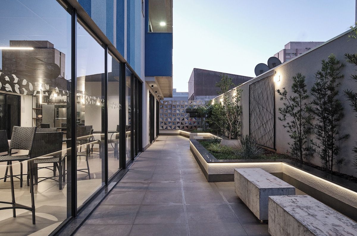 An outdoor seating area with modern furniture and a glass wall, surrounded by a landscaped garden and a building facade.