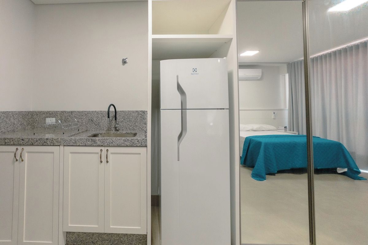 A compact hotel room with a kitchenette, a bed with a teal bedspread, and a glass partition separating the room from the adjacent space.