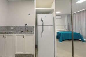 Une chambre d’hôtel compacte dotée d’une kitchenette, d’un lit recouvert d’une courtepointe turquoise et d’une cloison en verre qui sépare la chambre de l’espace adjacent.