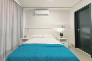 Quarto de hotel com cama, mesa de cabeceira, luminária, porta e ar condicionado.