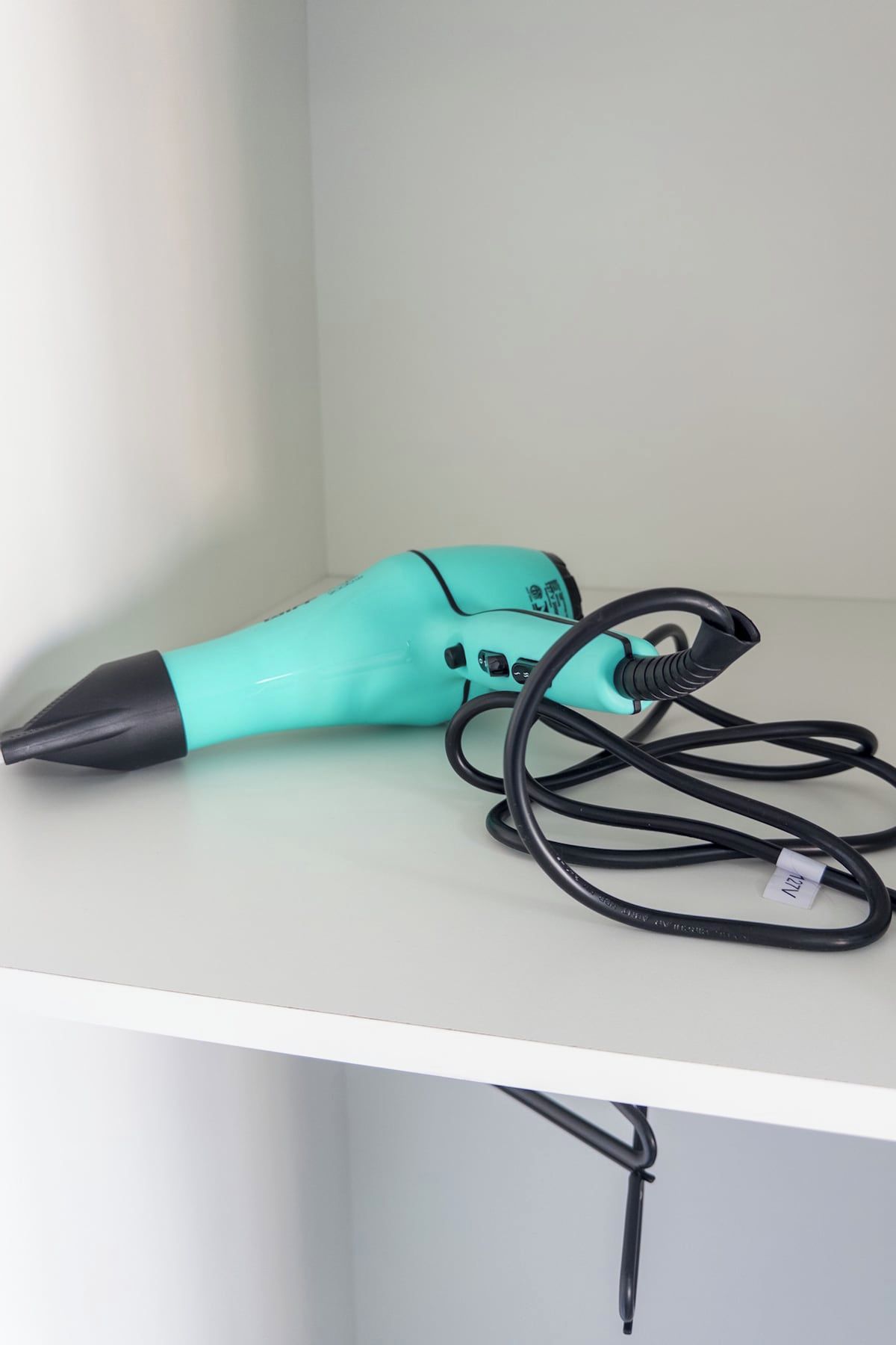 Un sèche-cheveux de couleur turquoise, doté d’un embout et d’un cordon noirs, est posé sur une étagère blanche.