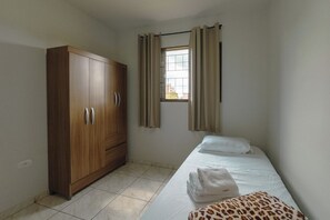 A single bed with a white mattress and two pillows, a wooden wardrobe, a window with curtains, and a tiled floor. - Budget Apartment near Unicesumar - GI08 (Maringá)