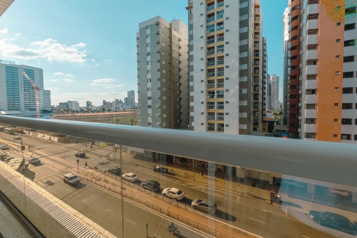 Um edifício alto com vários andares, céu claro e uma rua movimentada com carros e uma faixa de pedestres.