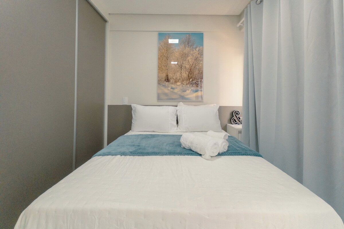 A hotel room with a bed, pillows, a blanket, a towel, a nightstand, a lamp, and a window with a view of trees.
