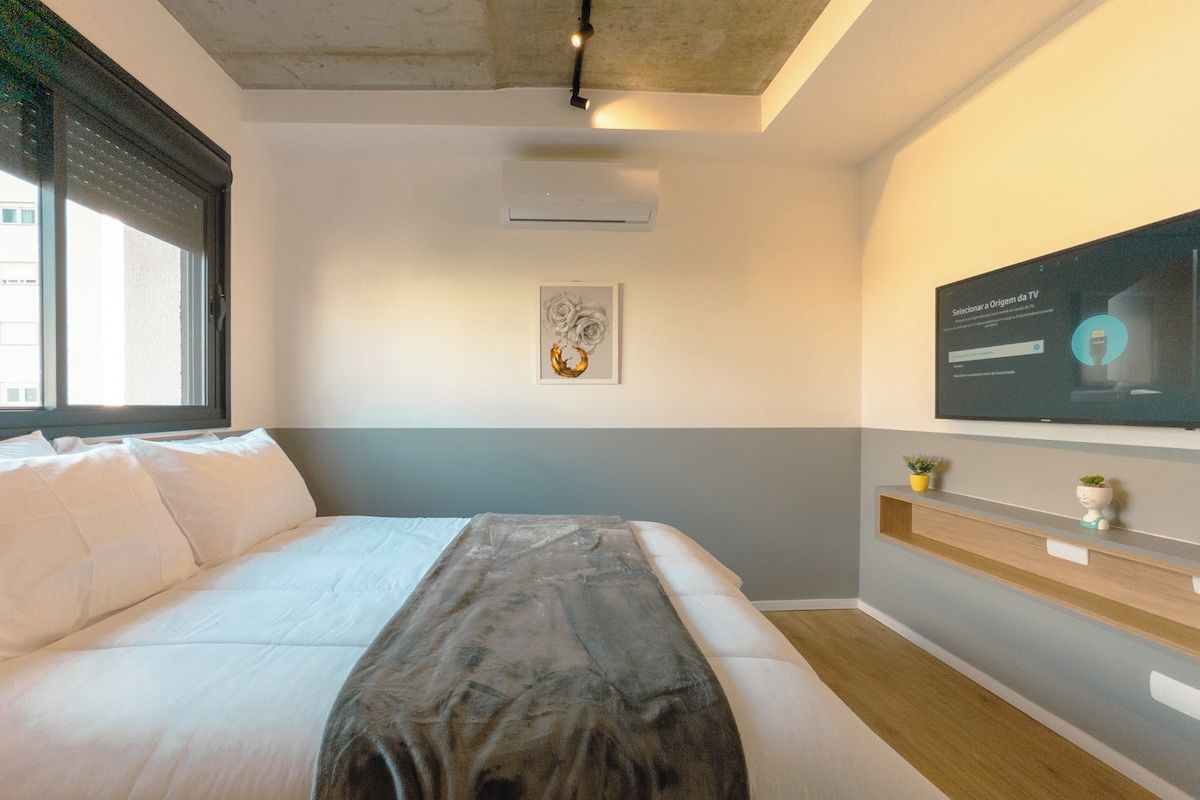 A modern hotel room with a bed, a mounted TV, a wall-mounted air conditioner, and a small shelf with decorative items.