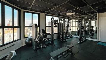 Uma academia com diversos equipamentos de exercício, janelas amplas e vista para a cidade.