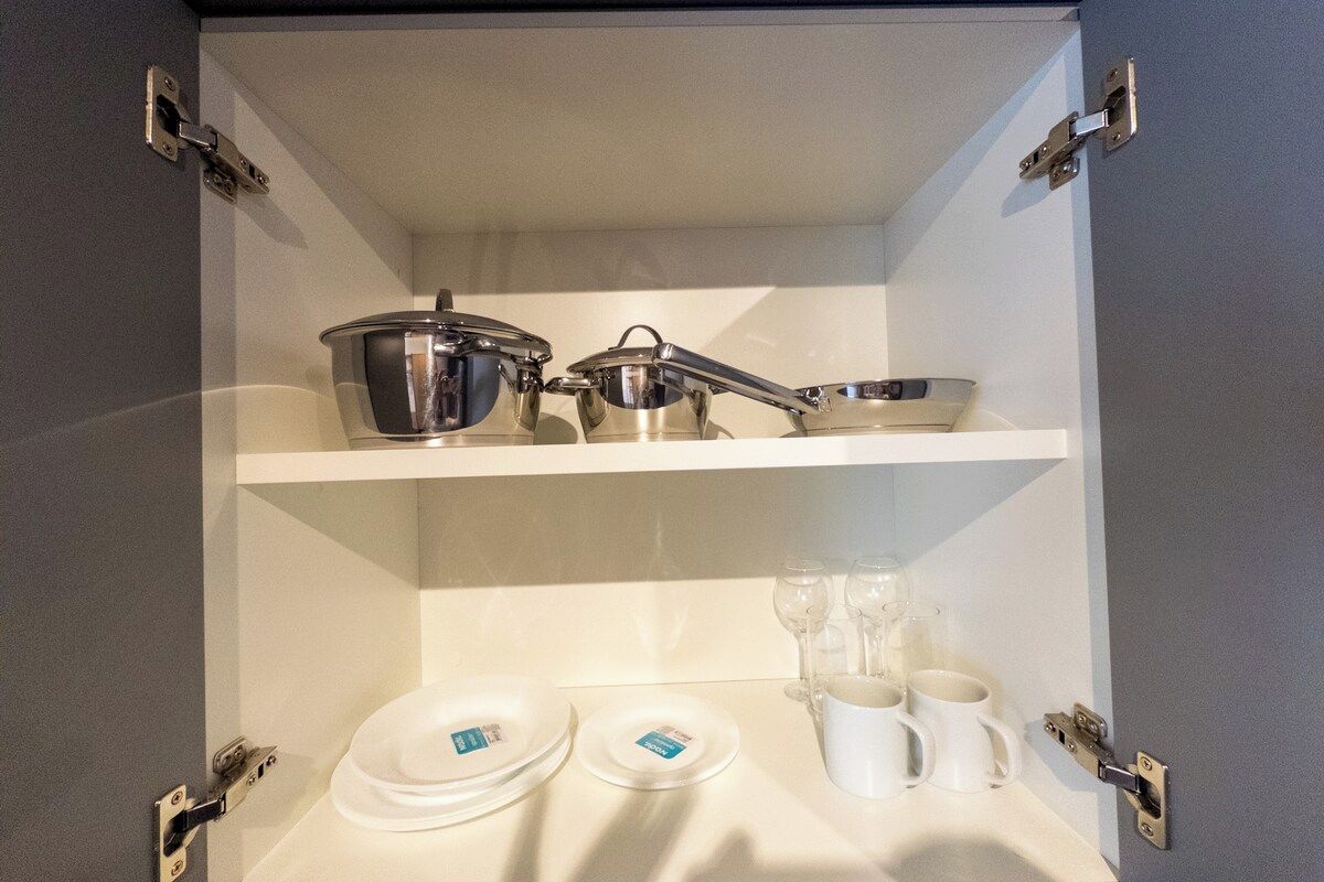 A kitchen cabinet with stainless steel cookware, a white shelf, and white plates and cups.