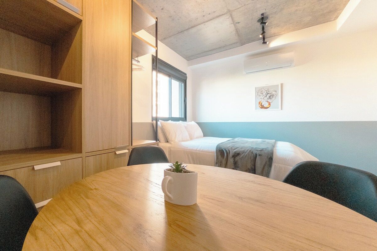 Una habitación moderna con una mesa de comedor de madera, una cama con ropa blanca, una ventana y una obra de arte colgada en la pared.