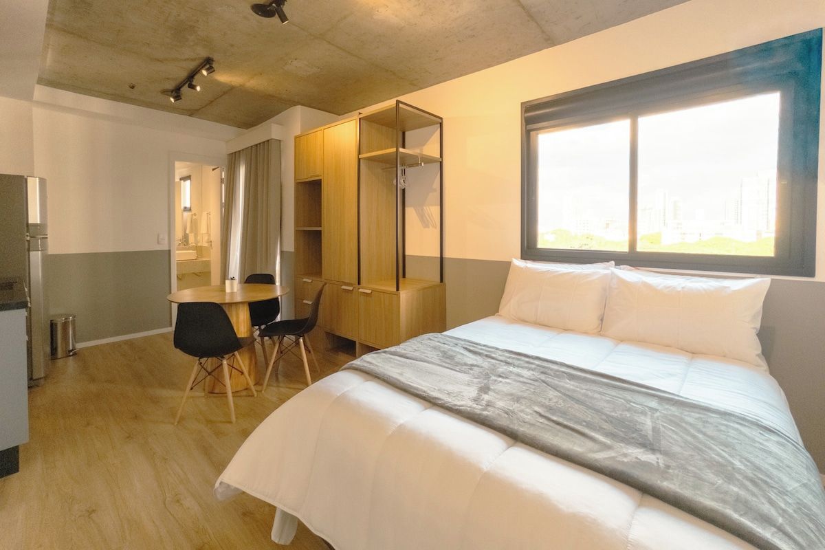Quarto de hotel com cama, escrivaninha, cadeira, guarda-roupa e janela.