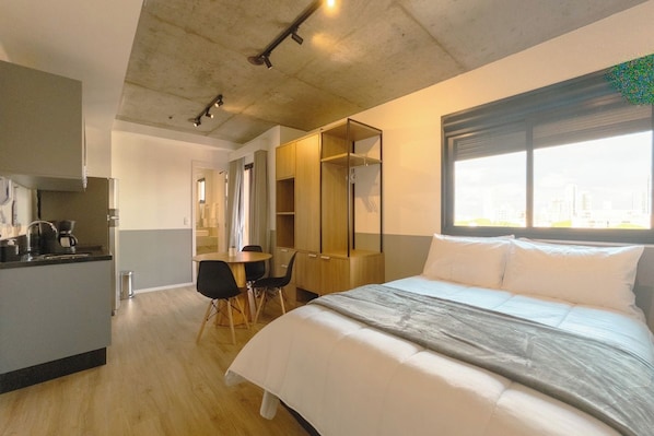 Quarto de hotel moderno com cama, escrivaninha e cozinha compacta.