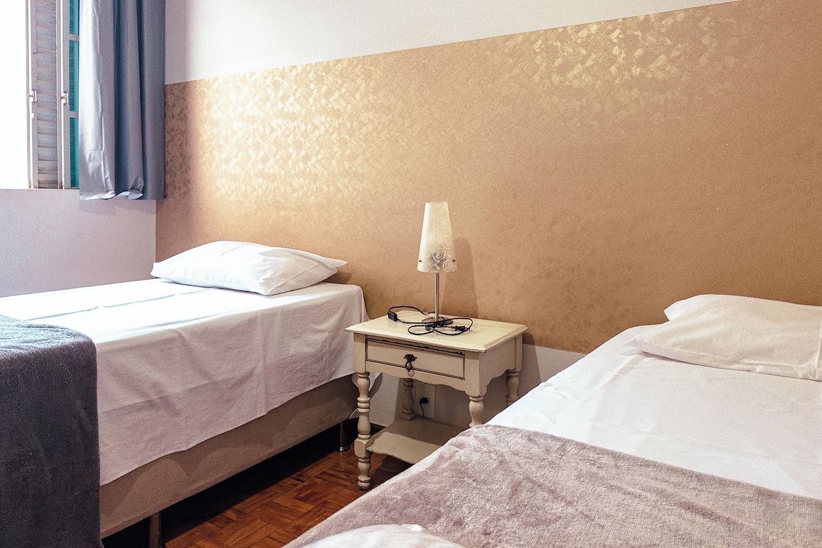 A hotel room with two beds, a bedside table with a lamp, and a window with curtains.