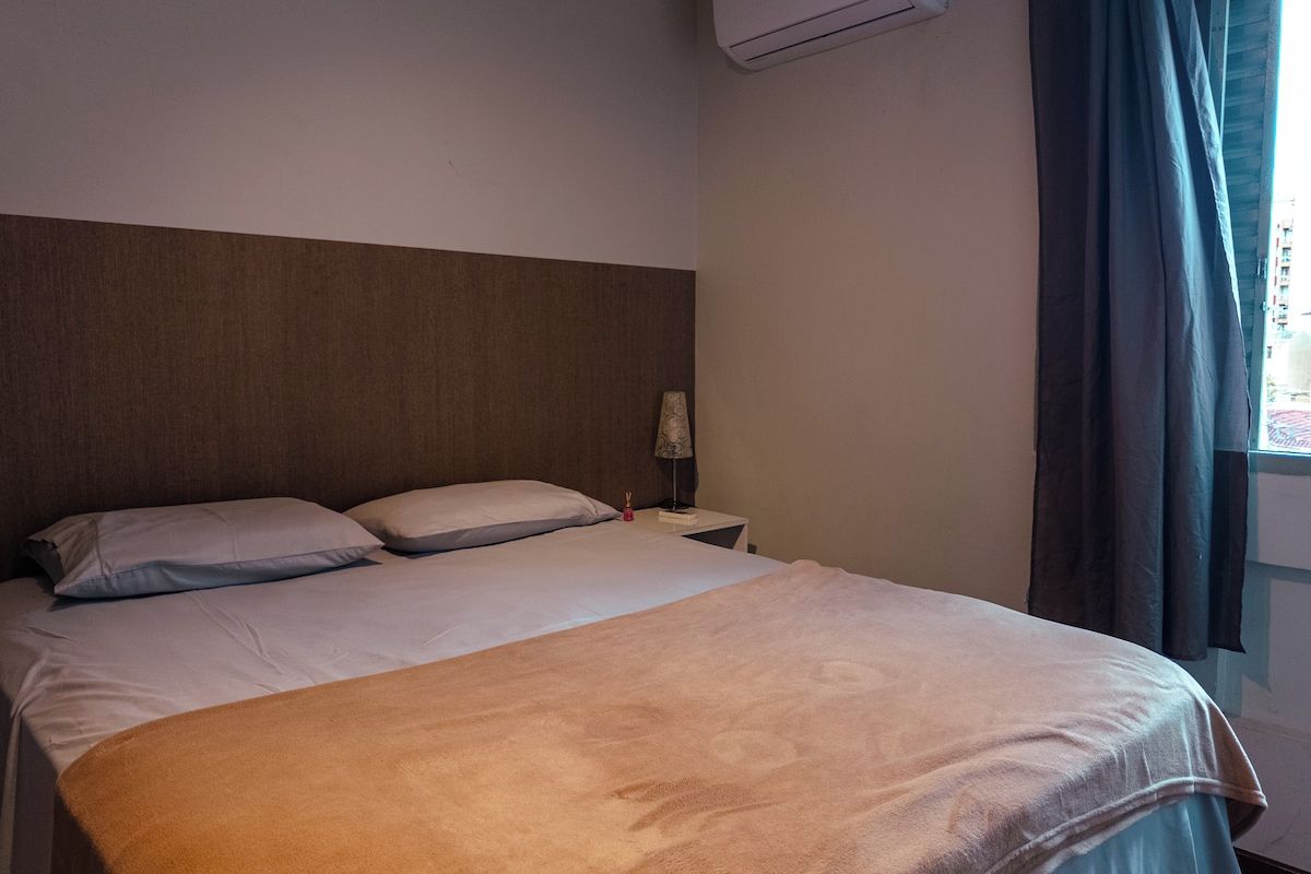 Quarto de hotel com cama, mesa de cabeceira, abajur e janela com cortinas.