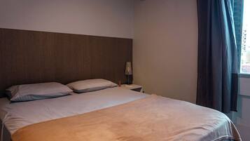 Quarto de hotel com cama, mesa de cabeceira, abajur e janela com cortinas.