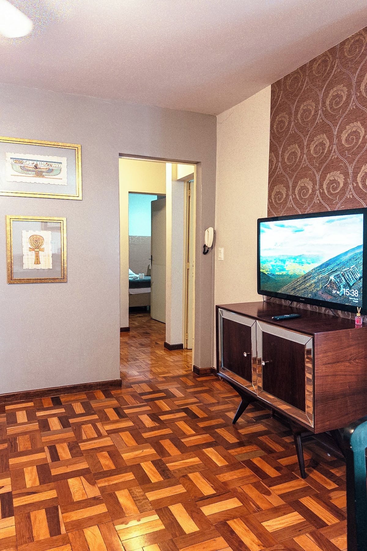 A living room with a wooden parquet floor, a television on a wooden stand, and framed pictures on the wall.