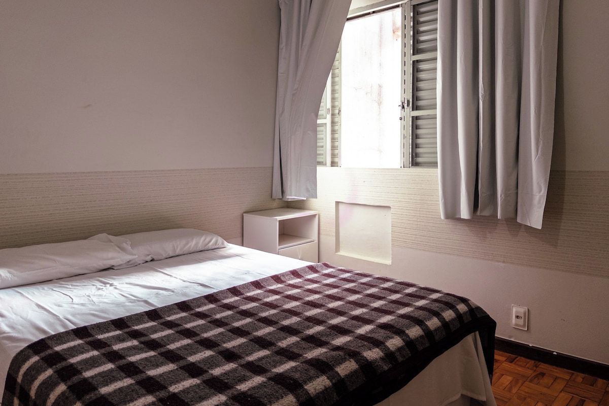 A bedroom with a bed, a bedside table, a window with curtains, and a checkered blanket.