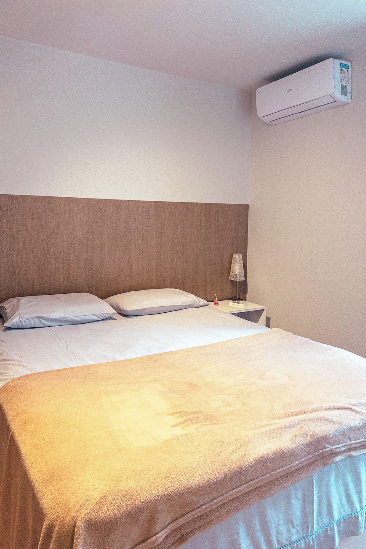 Quarto de hotel com cama, almofadas, mesa de cabeceira com abajur e ar condicionado na parede.