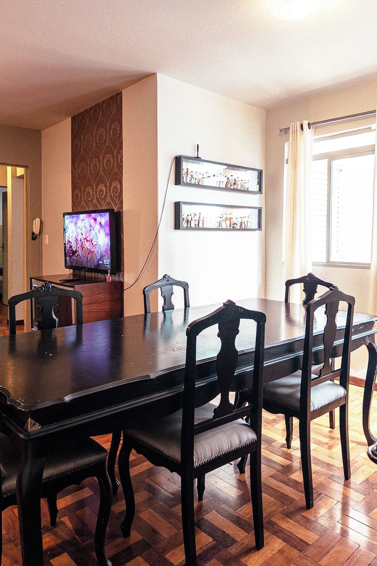 A dining area with a long table and six chairs, a television on a stand, and a wall-mounted shelf with bottles.