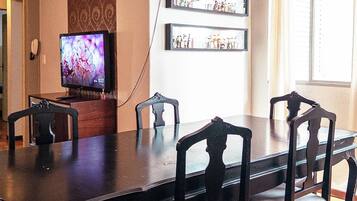 A dining area with a long table and six chairs, a television on a stand, and a wall-mounted shelf with bottles.