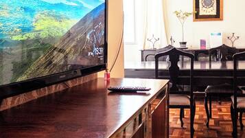 Quarto com televisão sobre um móvel de madeira, mesa de jantar com cadeiras e parede com padrão.