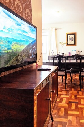 A room with a television on a wooden cabinet, a dining table with chairs, and a patterned wall. - Large 3-Bedroom Apartment near UEM - EL08 (Maringá)