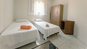 Quarto pequeno e limpo com duas camas de solteiro, um guarda-roupa de madeira, um ventilador de teto e uma janela com cortinas.