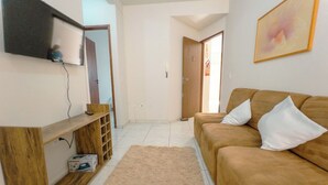 Uma sala de estar com um sofá, uma televisão presa à parede, uma estante de madeira e um tapete.