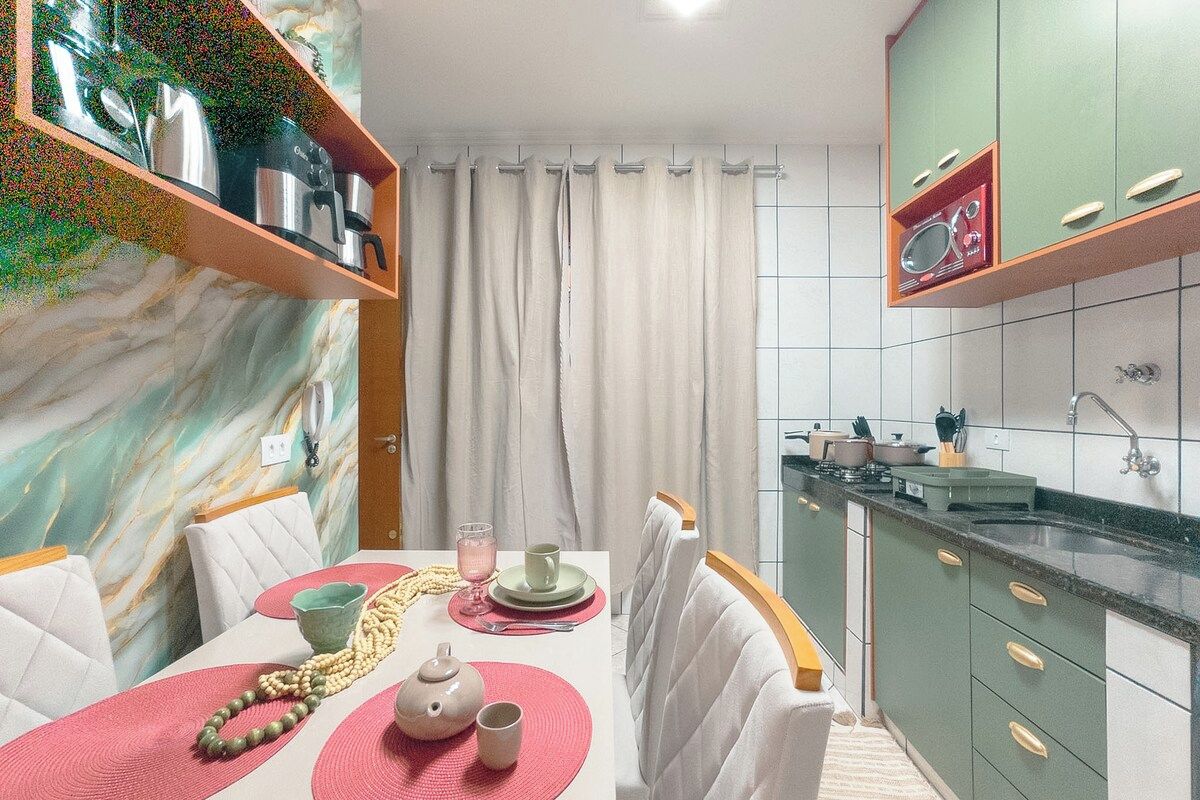 Cozinha moderna com área de refeições, apresentando painel de fundo de mármore, armários verdes e uma mesa de jantar com jogos americanos rosa e cadeiras brancas.