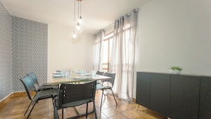 Uma área de refeições com uma mesa posta para quatro pessoas, cadeiras, um lustre e uma janela com cortinas.