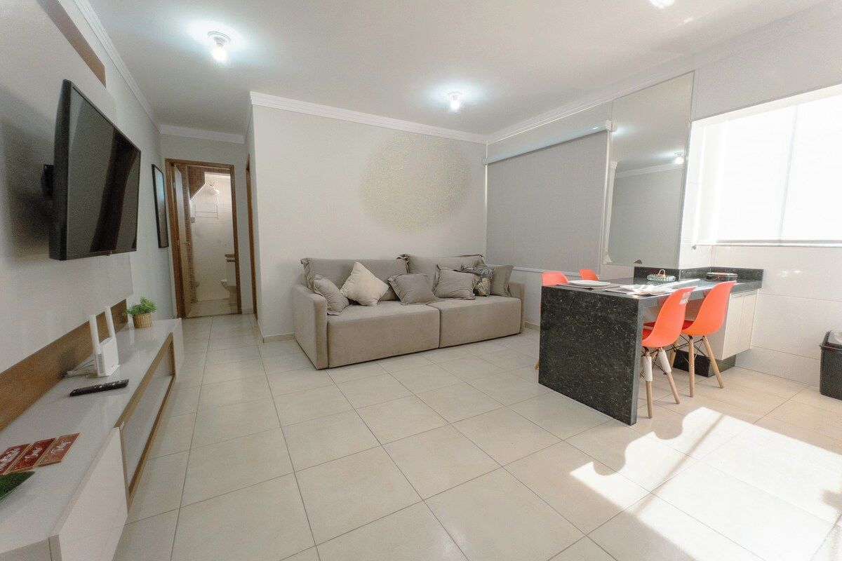 A modern living space with a sofa, a dining area with chairs, a flat-screen TV mounted on the wall, and a kitchen counter.