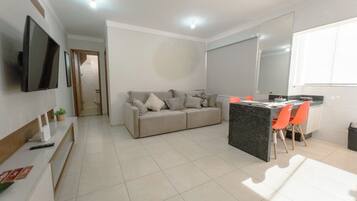 A modern living space with a sofa, a dining area with chairs, a flat-screen TV mounted on the wall, and a kitchen counter.