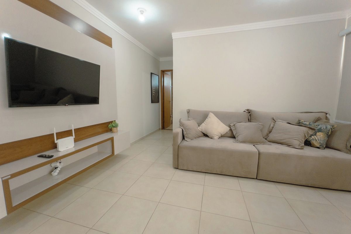 Ein Wohnzimmer mit einem an der Wand befestigten Flachbildfernseher, einem beige gefärbten Sofa mit vielen Kissen und einem Holztischchen mit einem weißen Gerät und einer kleinen Pflanze.