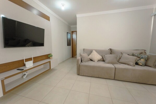 A living room with a flat-screen TV mounted on the wall, a beige sofa with multiple cushions, and a wooden console table with a white device and a small plant.