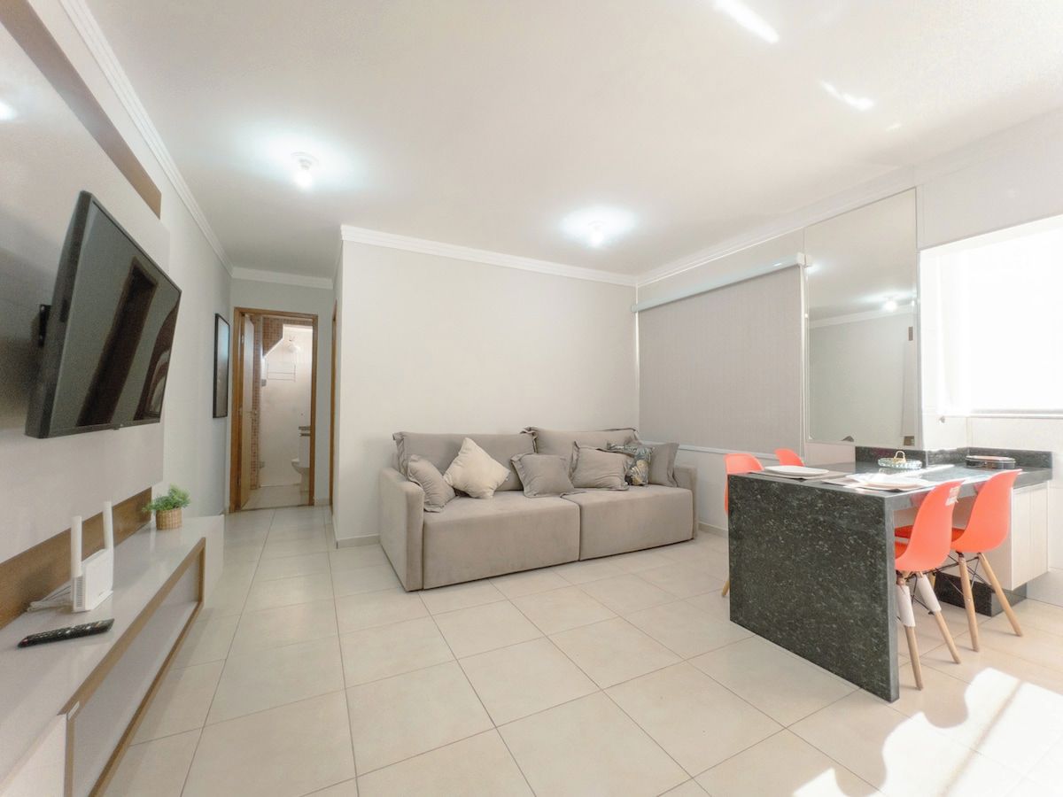 Una sala moderna con sofá, comedor con sillas, televisor montado en la pared y una cocina con mostrador.