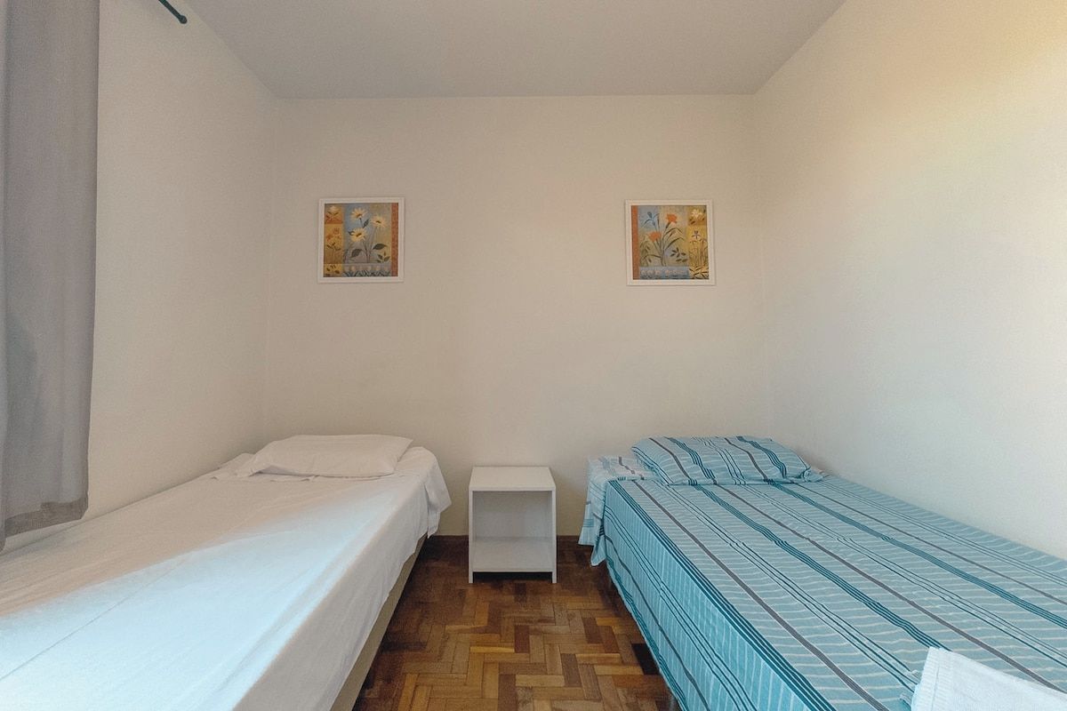 Acomodação compacta, com um quarto único, duas camas, uma mesa de cabeceira branca e duas pinturas penduradas na parede.