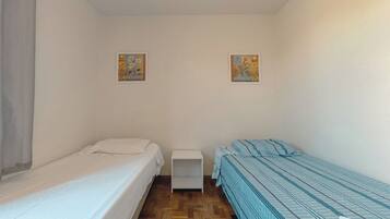 Acomodação compacta, com um quarto único, duas camas, uma mesa de cabeceira branca e duas pinturas penduradas na parede.