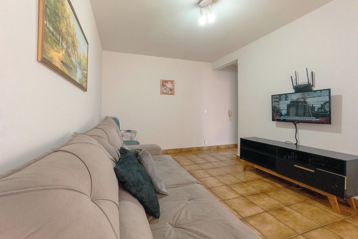Sala de estar com sofá bege, TV de tela plana na parede e rack de madeira para aparelhos eletrônicos.