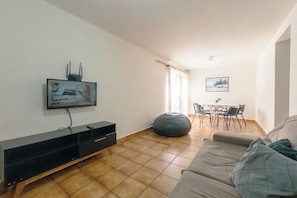 Una sala de estar con un televisor de pantalla plana montado en la pared, un sofá, un sillón de peluche, una mesa de comedor con sillas y una ventana con cortinas.