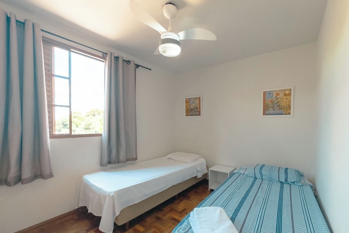 A small, single-room accommodation with a bed, a bedside table, a ceiling fan, and two wall-mounted pictures.