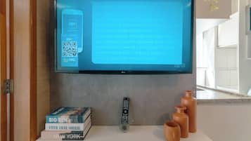 Seinälle asennettu litteänäyttöinen televisio, jonka päällä on QR-koodi ja tekstiä. Television alla on valkoinen hylly, jossa on pino kirjoja, kaukosäädin ja kolme keramiikkakuppia.