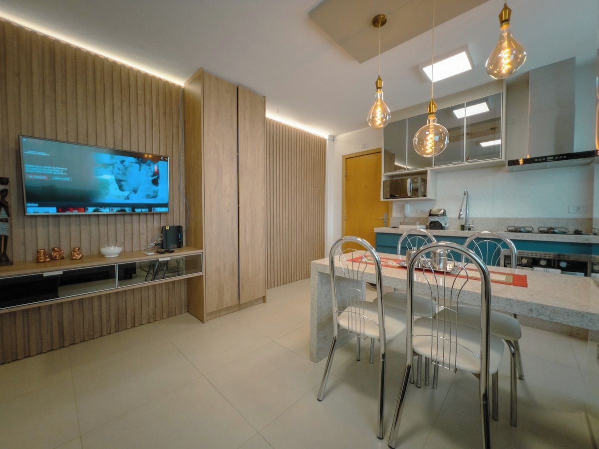 A modern kitchen with a built-in TV, dining area with chairs, and pendant lights.