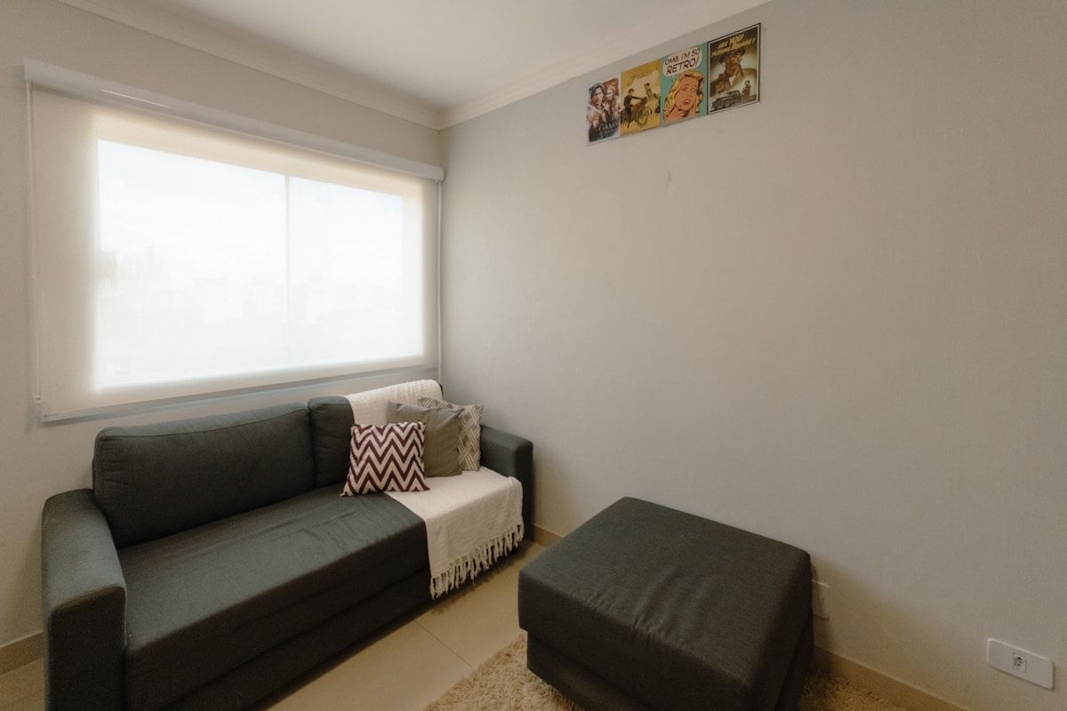 Una sala de estar con sofá, reposapiés y ventana con persianas.